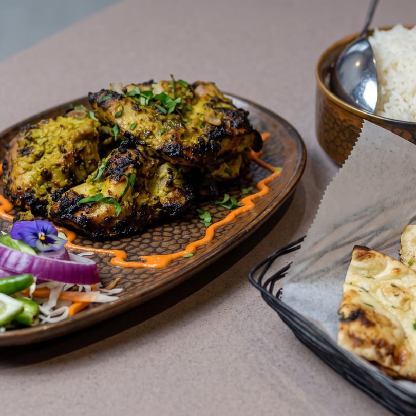 Grilled chicken tikka with rice and naan bread, garnished with red onion and a purple flower.