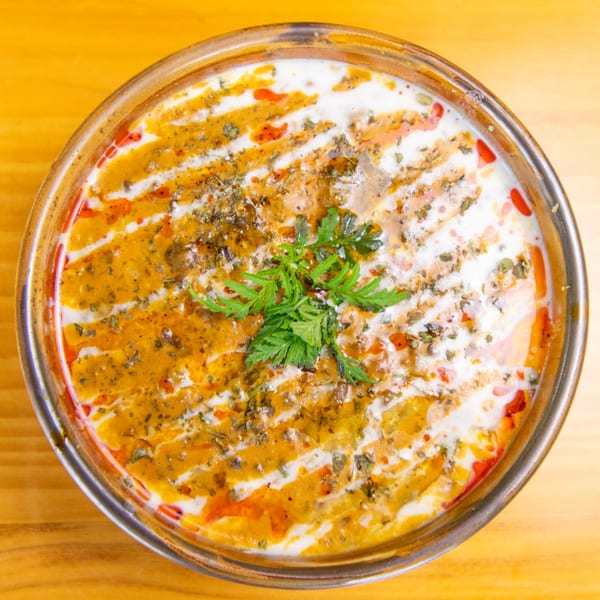 Overhead view of a bowl of creamy orange soup with white swirls, red oil, and green herbs.