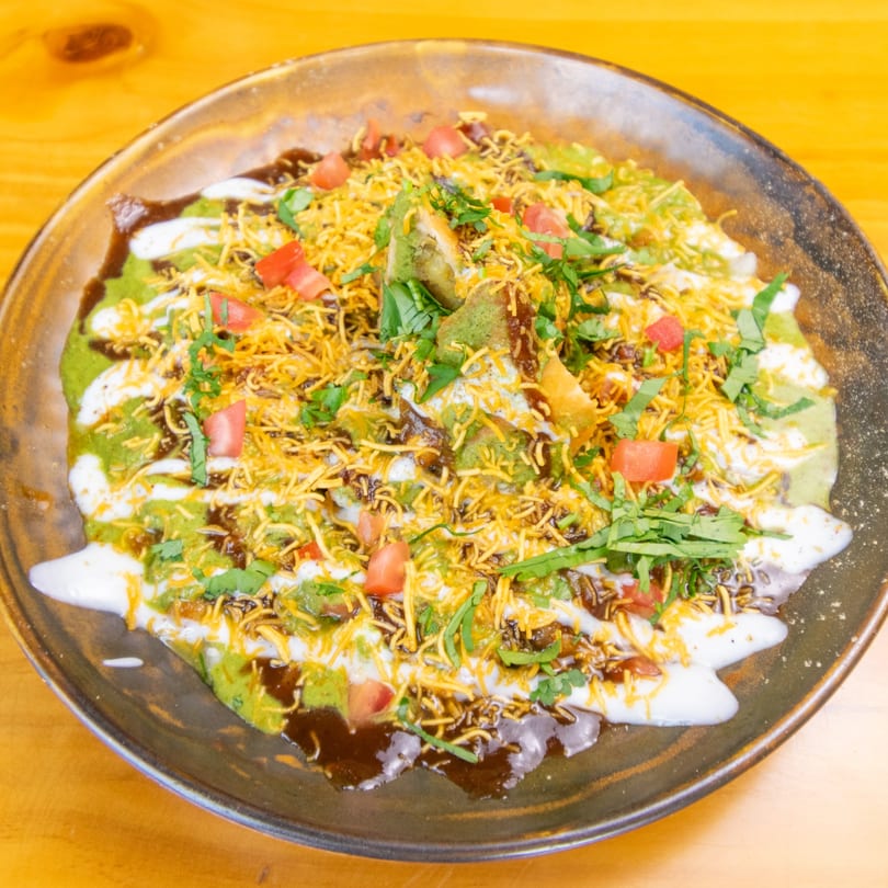 A vibrant plate of Indian chaat, topped with green chutney, yogurt, sev, and diced tomatoes.