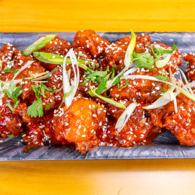 Close-up of crispy orange chicken with sesame seeds, green onions, and cilantro on a dark plate.