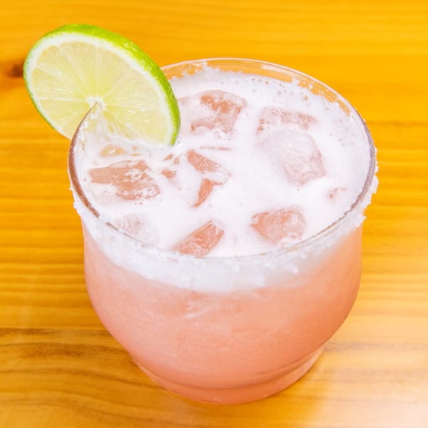 Pink cocktail with a foamy top, ice, and a lime wedge, on a wooden table.