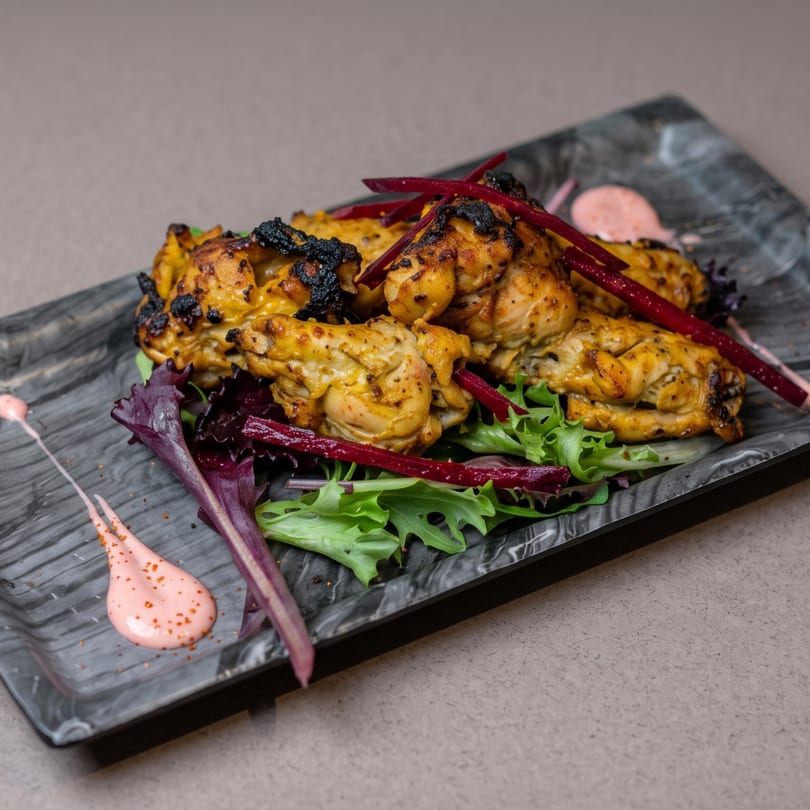 Grilled chicken pieces with charred edges, served on a bed of fresh greens with red accents and pink sauce.