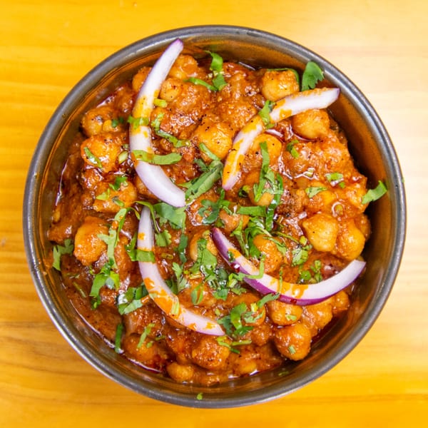 A close-up of chana masala, a chickpea curry, garnished with red onion and cilantro in a metal bowl.