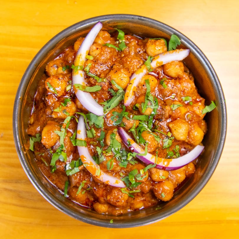 A close-up of chana masala, a chickpea curry, garnished with red onion and cilantro in a metal bowl.