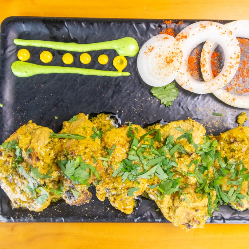 Chicken tikka with green sauce, yellow dots, and onion rings on a black plate.