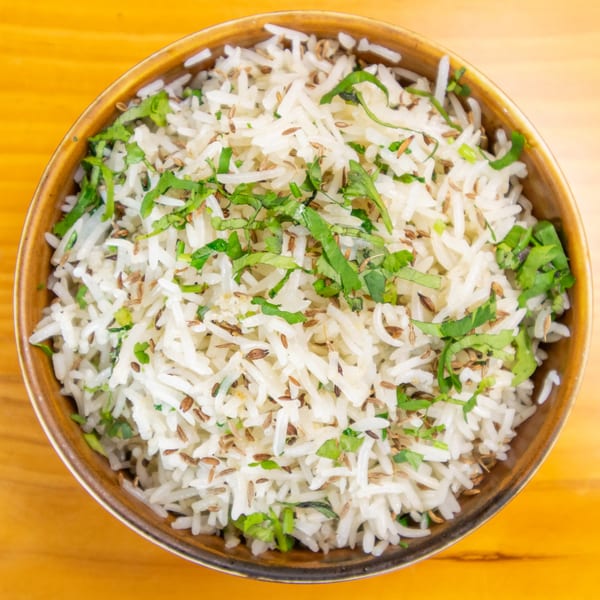 Overhead view of a brown bowl filled with white rice, green herbs, and cumin seeds.