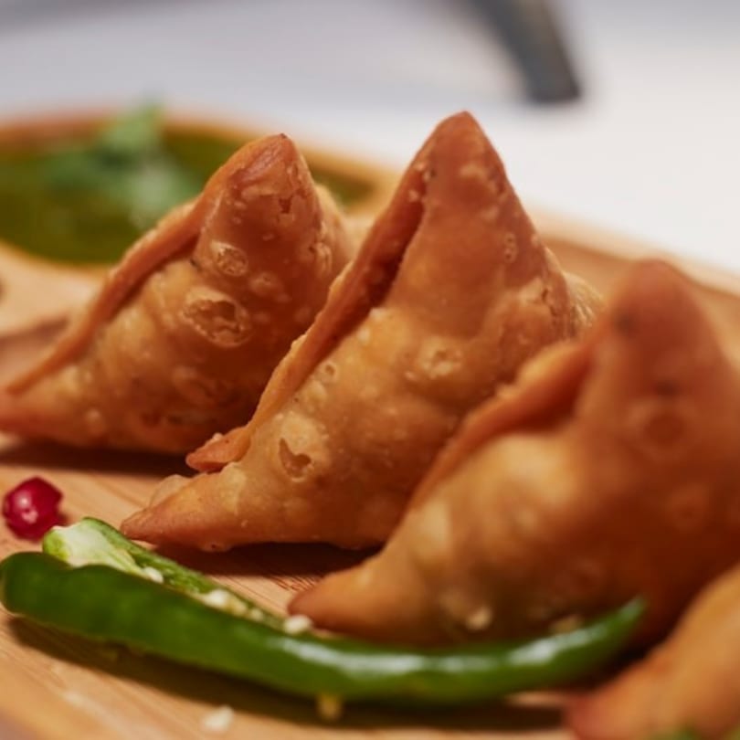 Three golden-brown samosas on a wooden board with green chili and cilantro chutney.