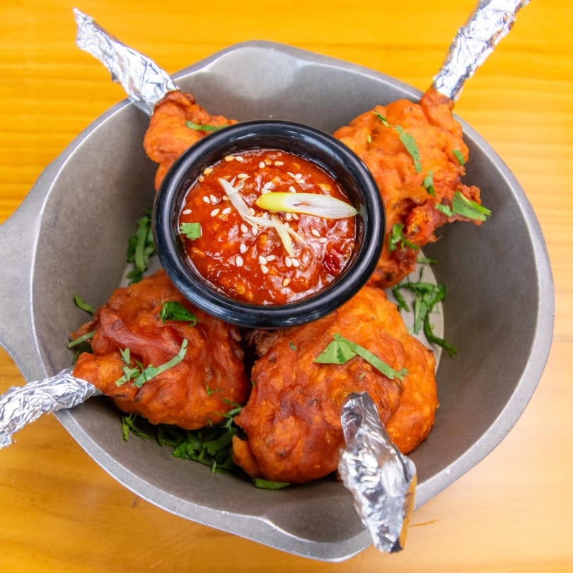 Four chicken lollipops with foil-wrapped bones, garnished with cilantro, and a side of red chili sauce.