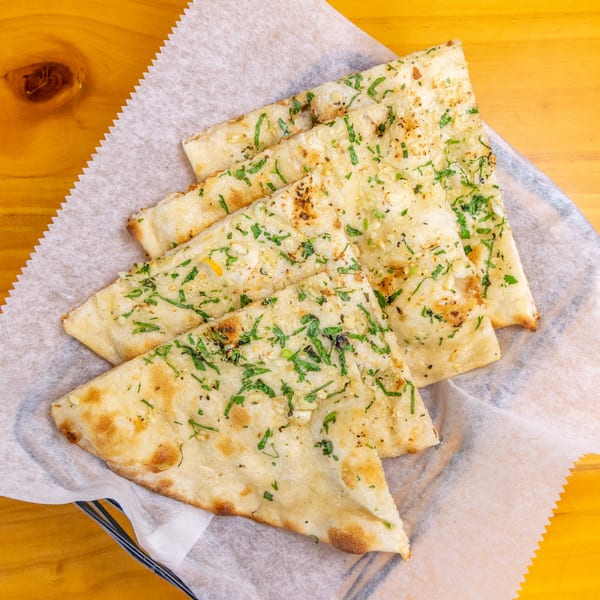 Four pieces of garlic naan bread, topped with green herbs, served in a basket with parchment paper.