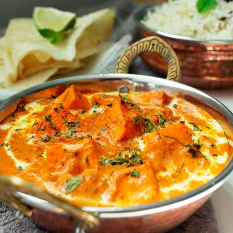 Indian butter chicken in a copper bowl with naan bread, lime, and rice in the background.