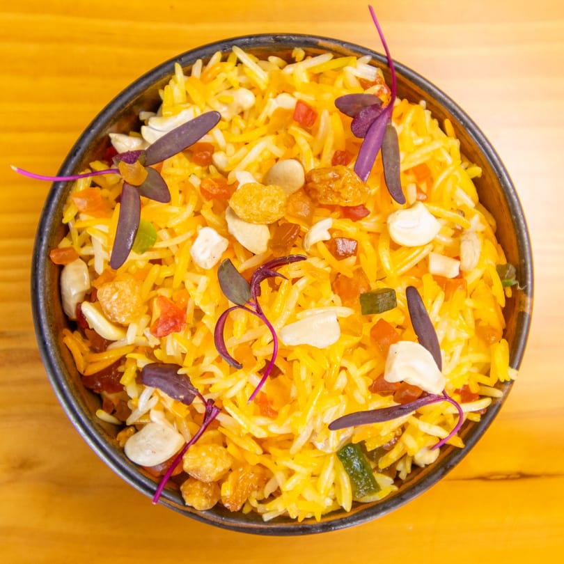 Overhead view of yellow rice with cashews, dried fruit, and purple garnish in a black bowl.