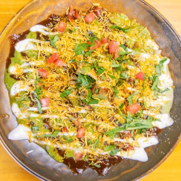 A vibrant Indian dish with green and white sauces, topped with golden crispy noodles, tomatoes, and cilantro.
