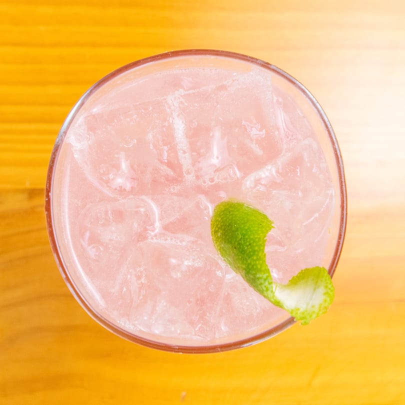 Overhead view of a pink cocktail with ice and a lime peel garnish on a wooden table.