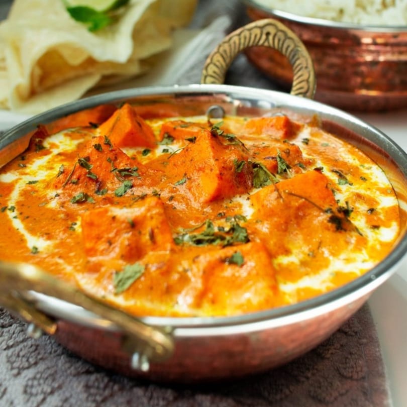 Indian butter chicken or paneer in a copper bowl, garnished with fresh herbs, with naan and rice.