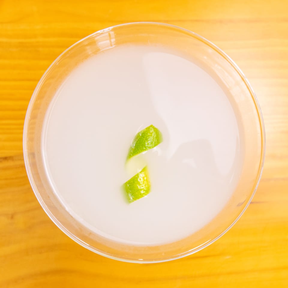 Overhead view of a white cocktail in a martini glass, garnished with a lime peel.