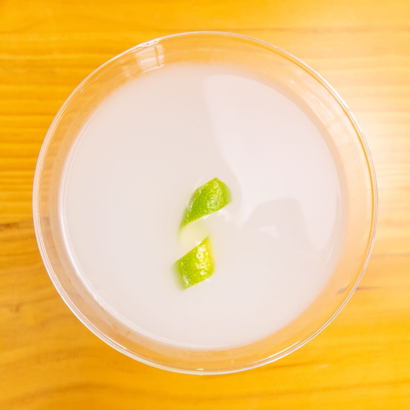 Overhead view of a white cocktail in a martini glass, garnished with a lime peel.