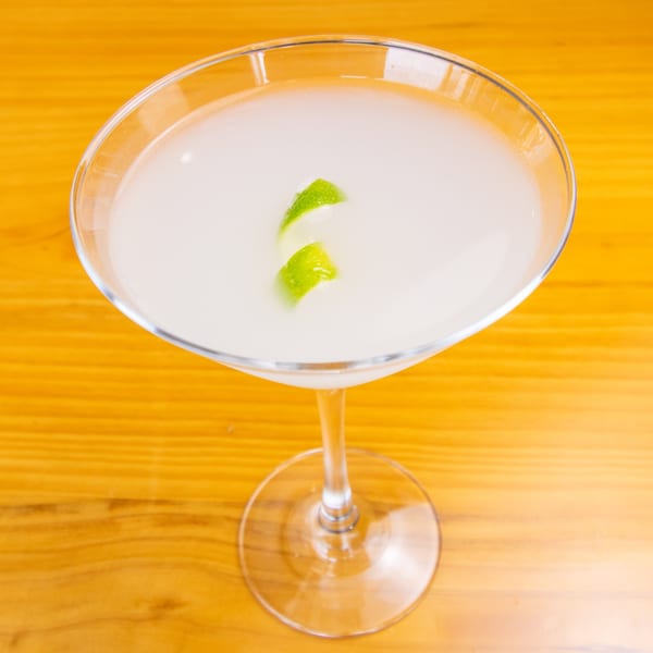 A light-colored cocktail in a martini glass, garnished with two lime peels, on a wooden table.