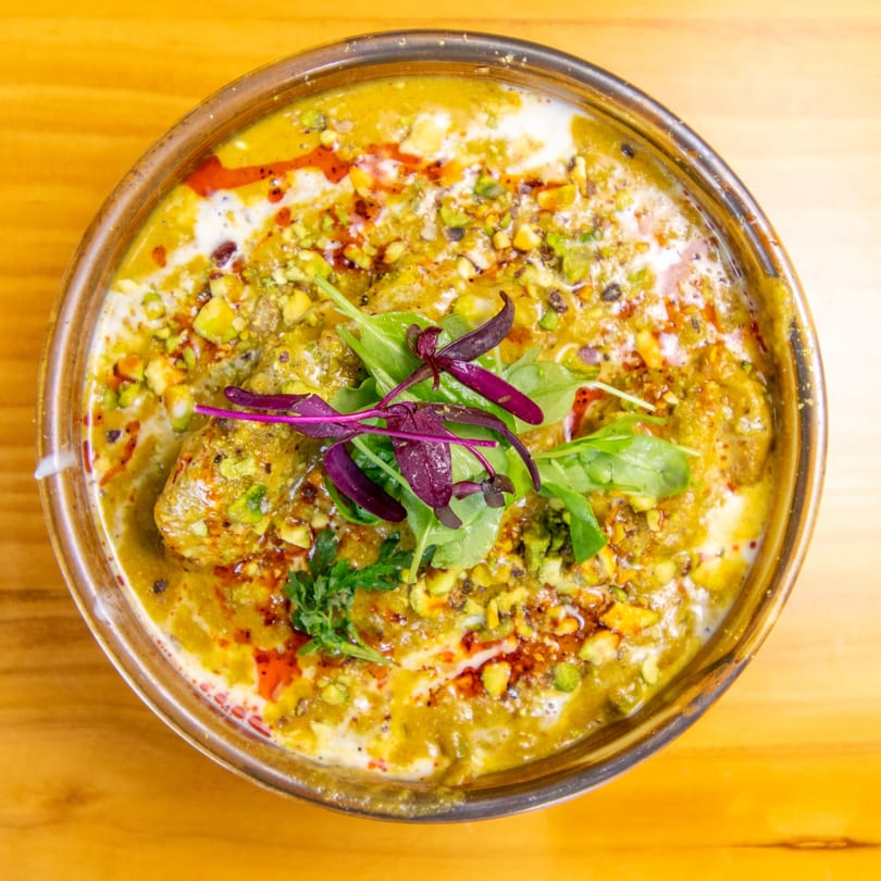 Overhead view of a vibrant green curry dish with cream, red oil, pistachios, and fresh herbs.